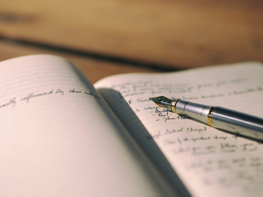 A pen rests in the seam of a notebook with handwritten text out of focus.